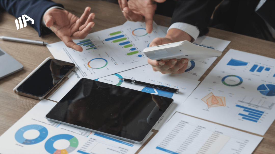 A workspace with several business documents, charts, and graphs spread across the table. Two individuals are involved in a discussion, using a calculator and pointing at the papers.