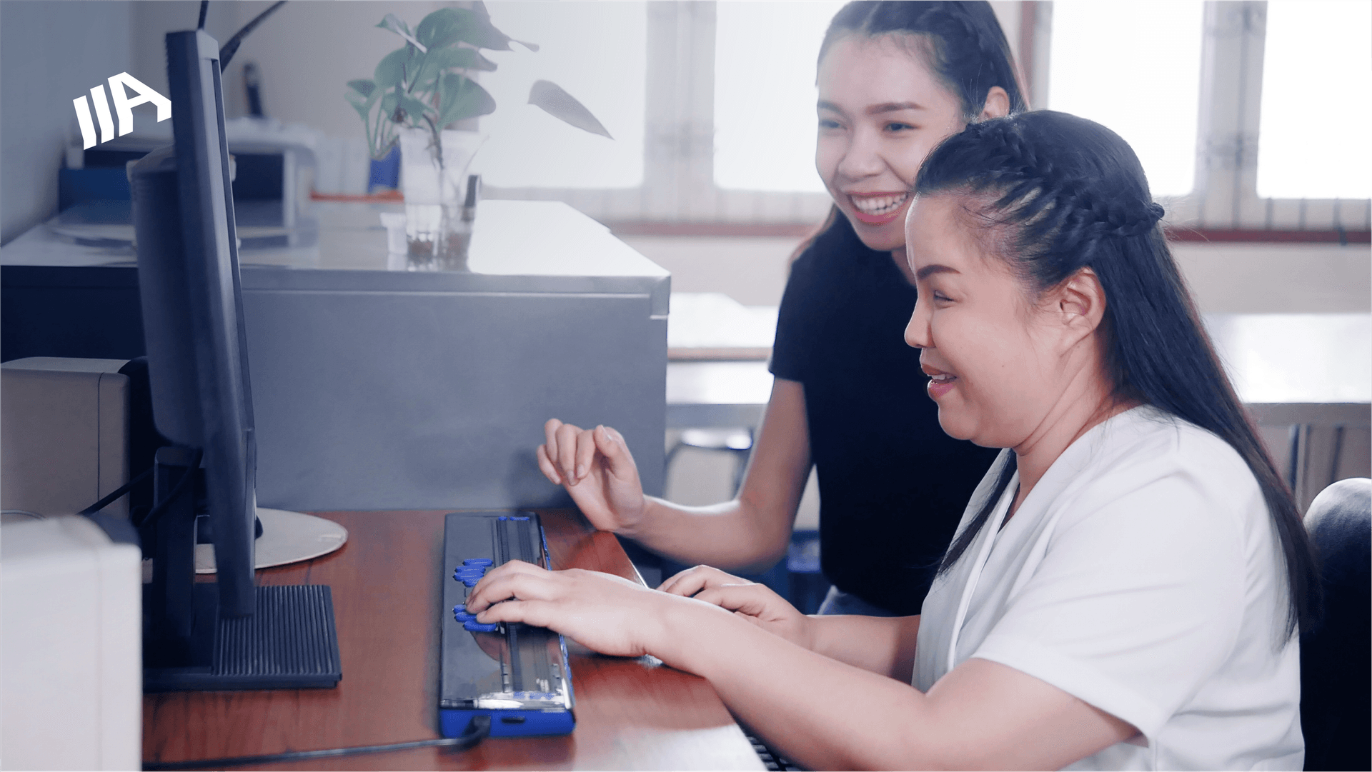 A visually impaired woman using a braille keyboard while working on a computer, assisted by a smiling colleague in an office setting.