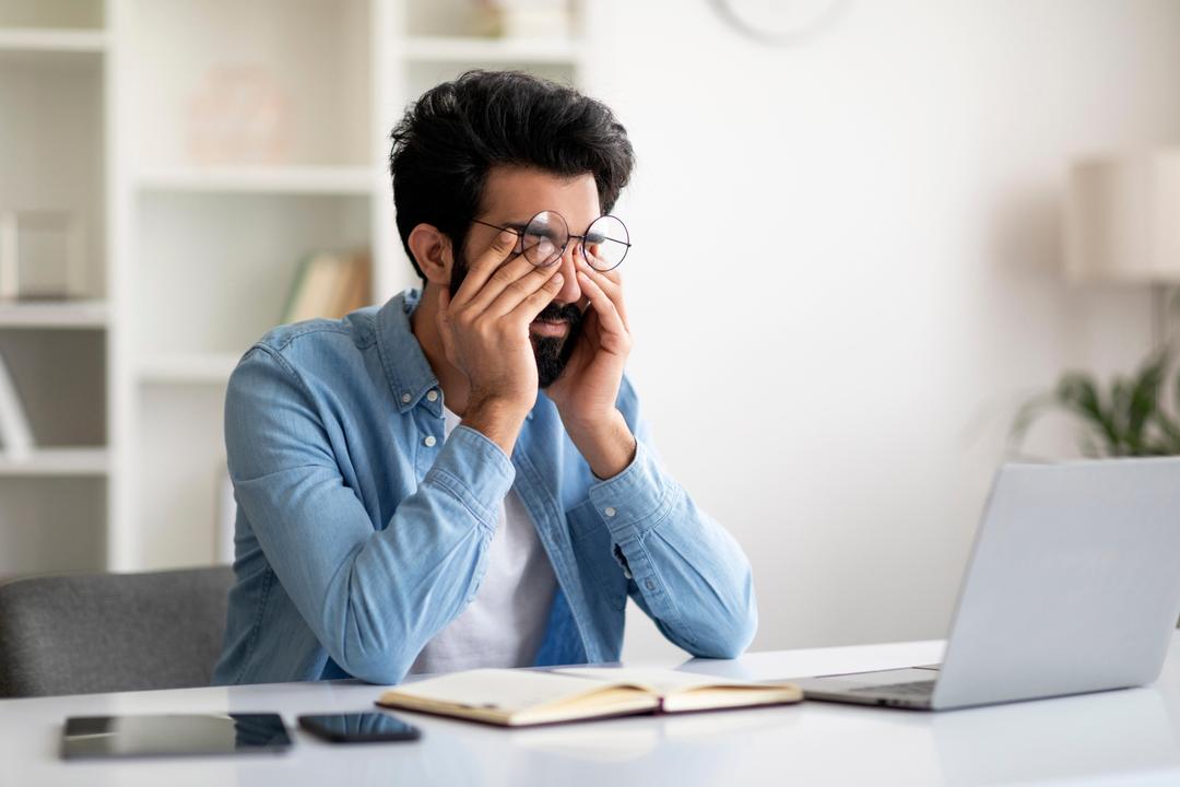 Man rubbing his tired eyes while sitting at a desk with a laptop